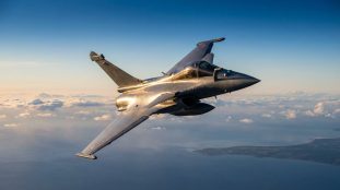 Chasseur Rafale français en plein vol, survolant la mer et la côte sous un ciel clair avec des nuages. L'avion brille au soleil.