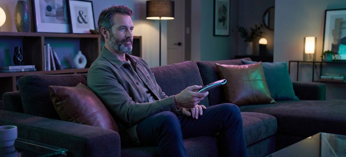 Homme barbu d'âge moyen sur un canapé sombre, télécommande en main, regardant la télévision dans un salon éclairé d'ambiance.