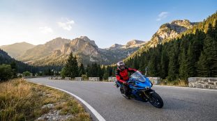Un motard en équipement complet conduit une moto bleue sur une route sinueuse au milieu des montagnes.