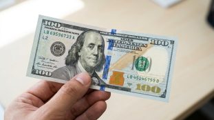 Une main tenant un nouveau billet de 100 dollars américain sur un fond de bureau flou.