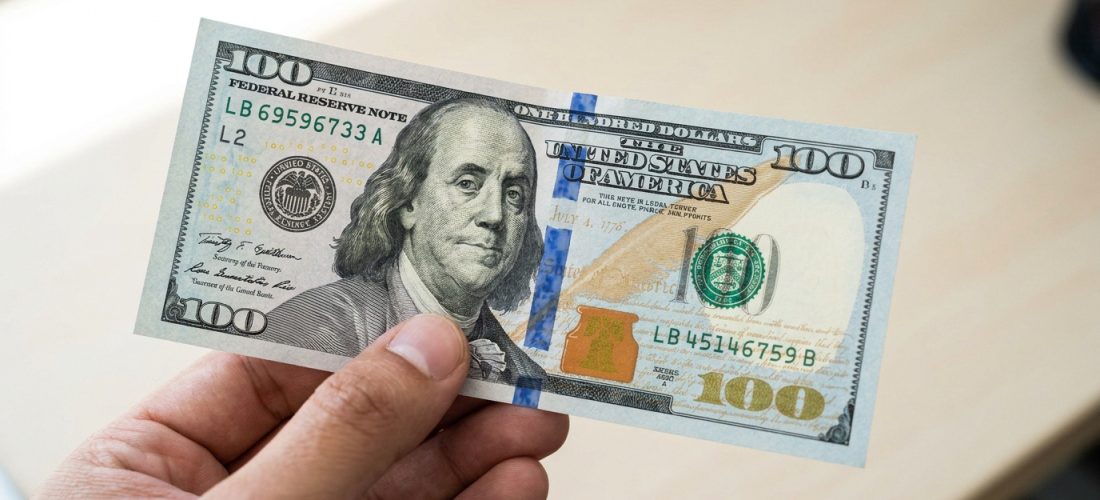 Une main tenant un nouveau billet de 100 dollars américain sur un fond de bureau flou.